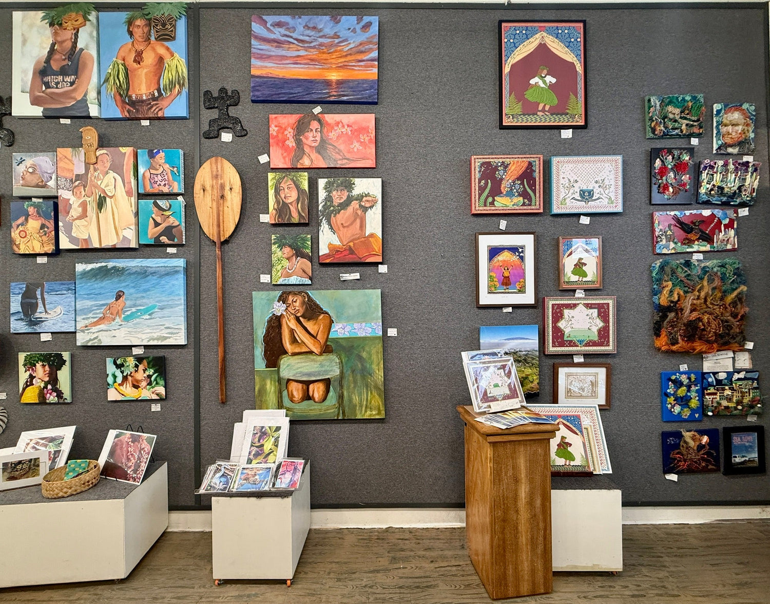 A gallery-style display of many colorful paintings and art prints mounted on a dark gray wall, with a few framed pieces and small books displayed on white pedestals in a gallery-like room.