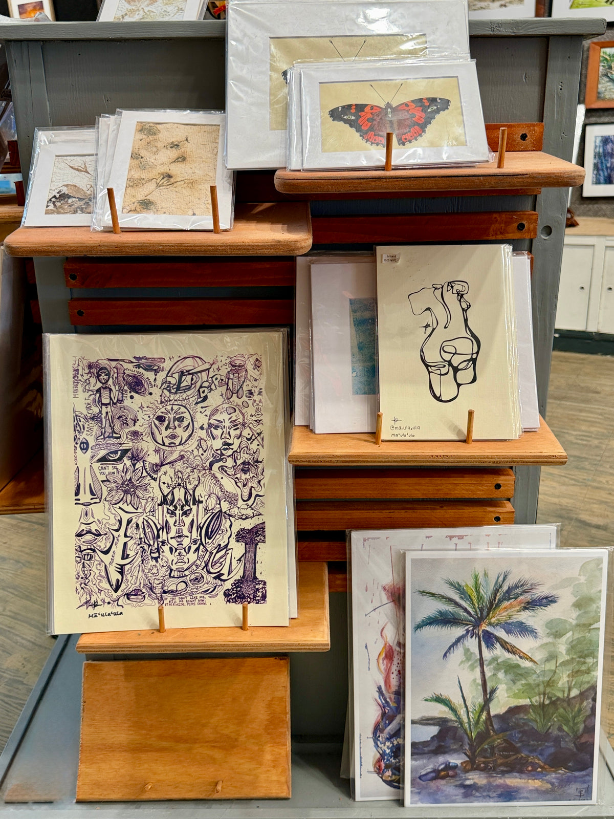 A display of framed and unframed art prints on wooden stands in a gallery or shop setting. The prints include a butterfly illustration, a purple line-art collage, a black line drawing of a face, a tropical landscape with palm trees, and other abstract or scenic prints.
