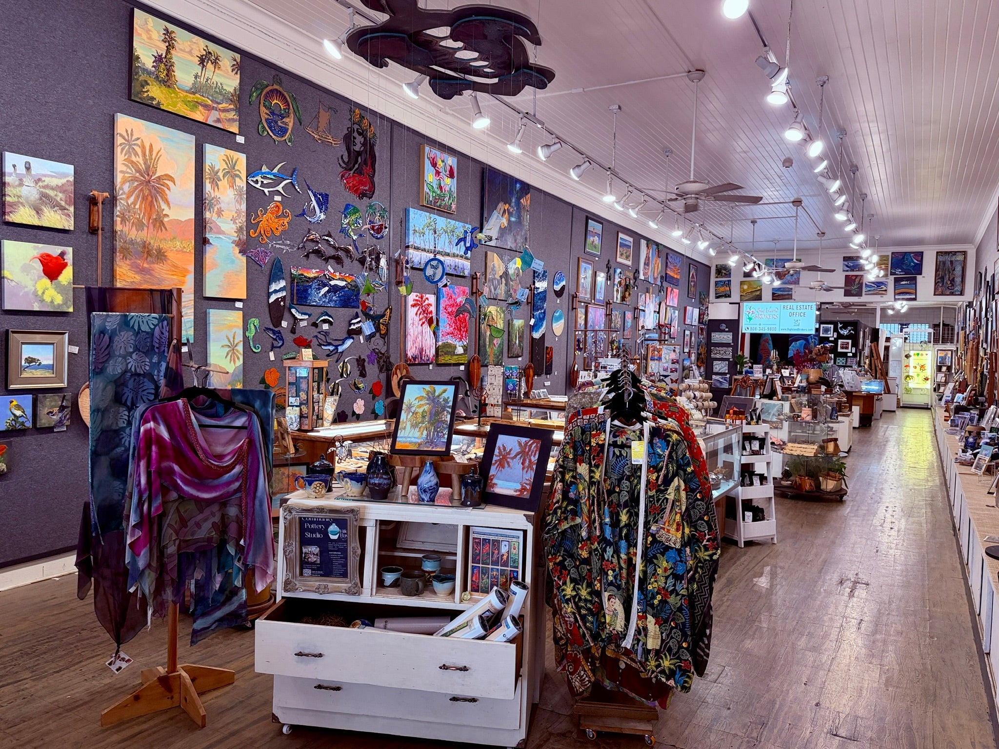 A wide view of One Gallery interior with colorful artwork, clothing on display stands, framed art, and merchandise arranged along a wooden floor aisle.