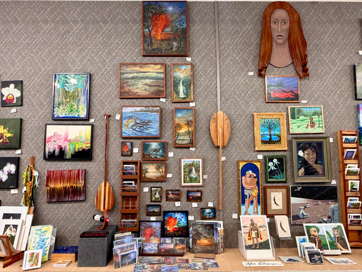 A gallery-style wall display of many framed and unframed paintings, prints, and small art pieces mounted on a gray patterned wall, with a large red-haired female bust sculpture above and wooden paddle-like decor hanging vertically.