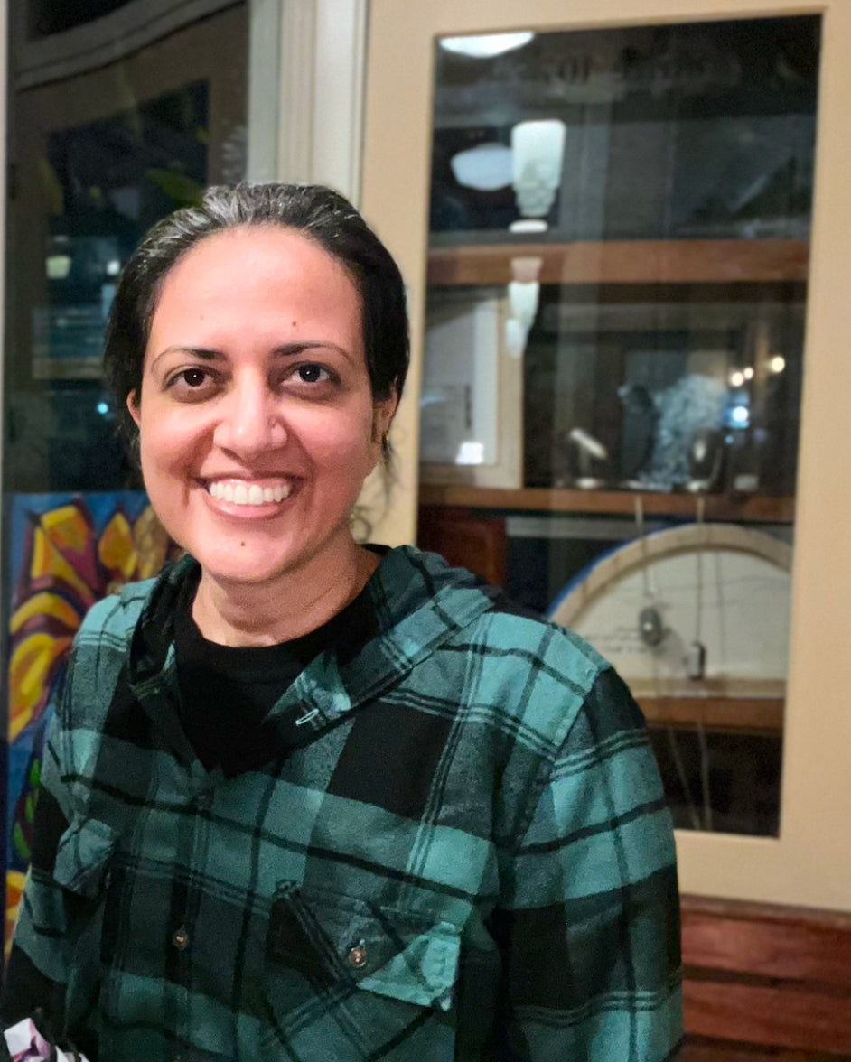 A smiling woman with dark hair pulled back, wearing a green and black plaid flannel shirt over a black top, standing in front of a glass display cabinet in an indoor setting.