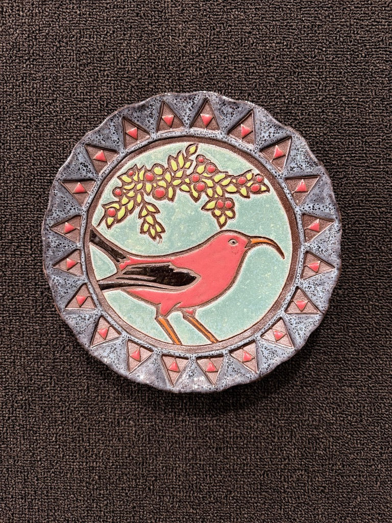 A round decorative ceramic plate or coaster featuring a red bird perched under a leafy branch with red berries, set against a textured dark brown background.