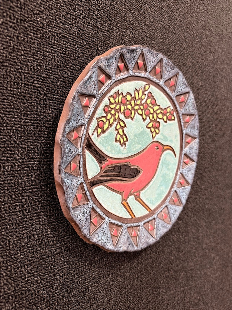 An oval decorative pin or badge featuring a red bird under a yellow flowering branch, set against a pale green background with a silver-gray patterned border, photographed on a dark brown textured fabric.