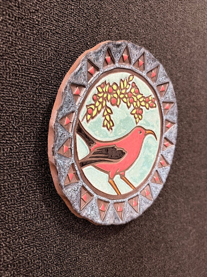 An oval decorative pin or badge featuring a red bird under a yellow flowering branch, set against a pale green background with a silver-gray patterned border, photographed on a dark brown textured fabric.