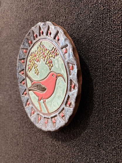 An oval decorative wall plaque featuring a carved bird design with a red bird, green background, and brown textured border, shown on a dark brown textured surface.
