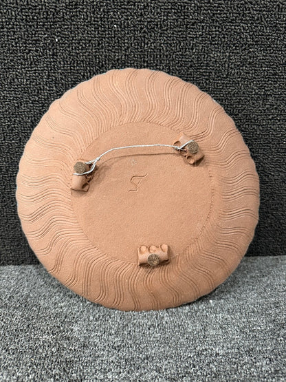 A round terracotta-colored wall hanging plaque with a carved sunburst pattern, shown from the back with metal hanging hardware against a dark textured backdrop.