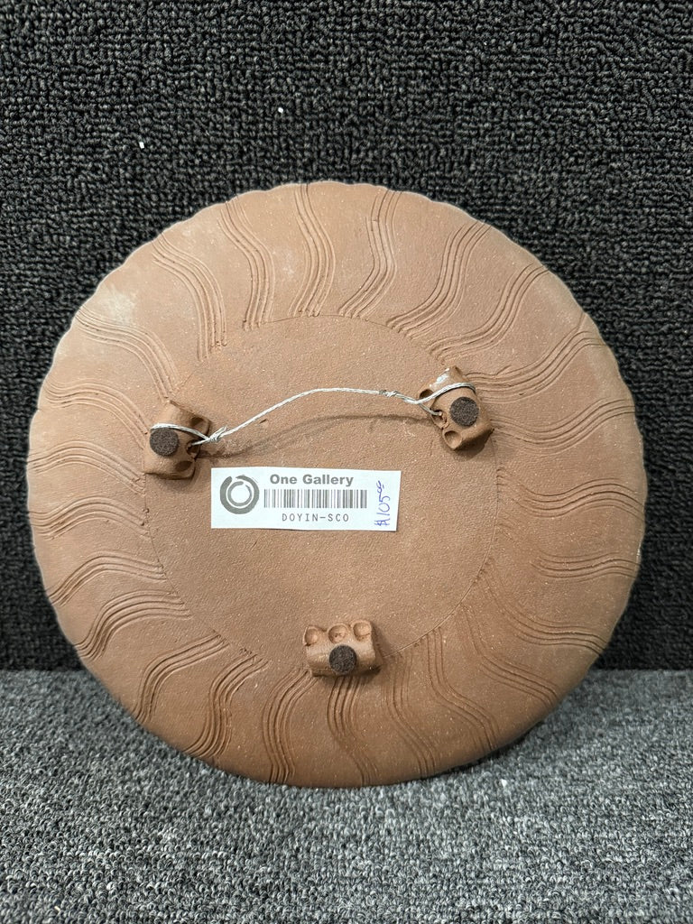 The back of a round, light brown ceramic plate or decorative tile, featuring a textured wavy pattern, a mounting wire with three small metal clips, and a white label reading 'One Gallery'.