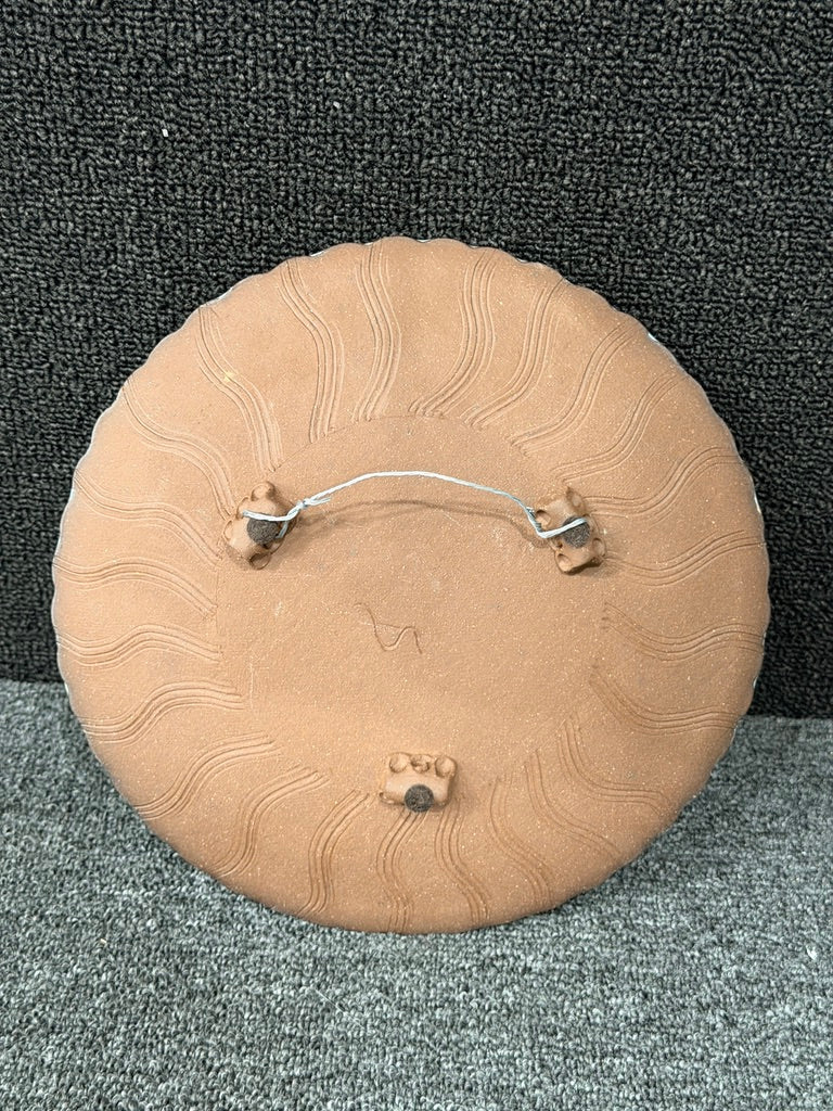 The back of a round terracotta-colored decorative plaque or medallion, featuring a wavy sunburst pattern and a metal wire hanger with two small hooks, shown on a textured gray and black background.