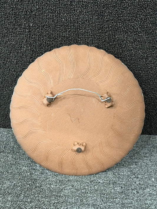 The back of a round terracotta-colored decorative plaque or medallion, featuring a wavy sunburst pattern and a metal wire hanger with two small hooks, shown on a textured gray and black background.