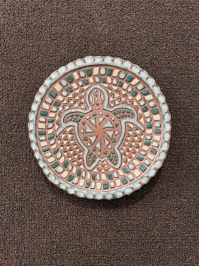 A round decorative plaque or medallion featuring an embossed sea turtle design with geometric patterns, finished in a warm metallic copper tone with small teal accents and a white beaded border, shown on a dark brown textured background.