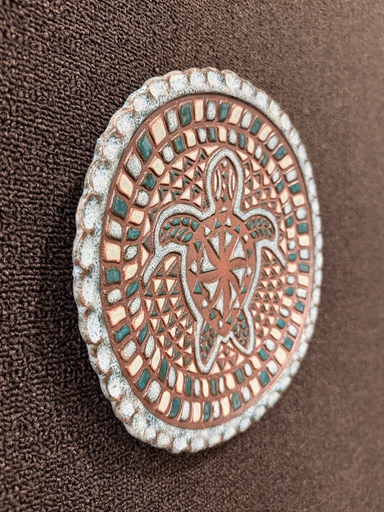 A close-up of a round decorative ceramic hanging plate featuring a raised sea turtle design, with a copper-toned center, silver-toned border, and small teal accents, set against a dark brown textured fabric background.