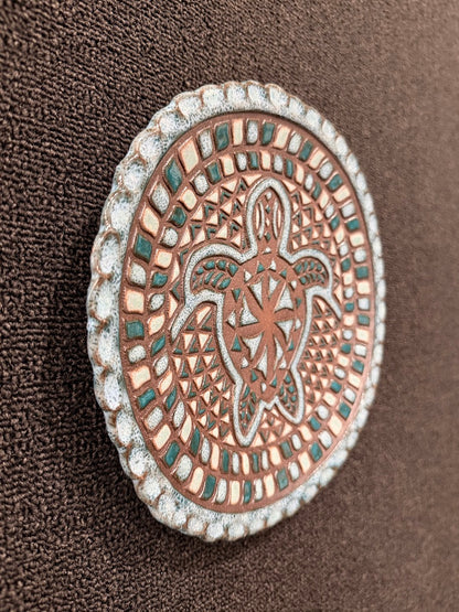 A close-up of a round decorative ceramic hanging plate featuring a raised sea turtle design, with a copper-toned center, silver-toned border, and small teal accents, set against a dark brown textured fabric background.