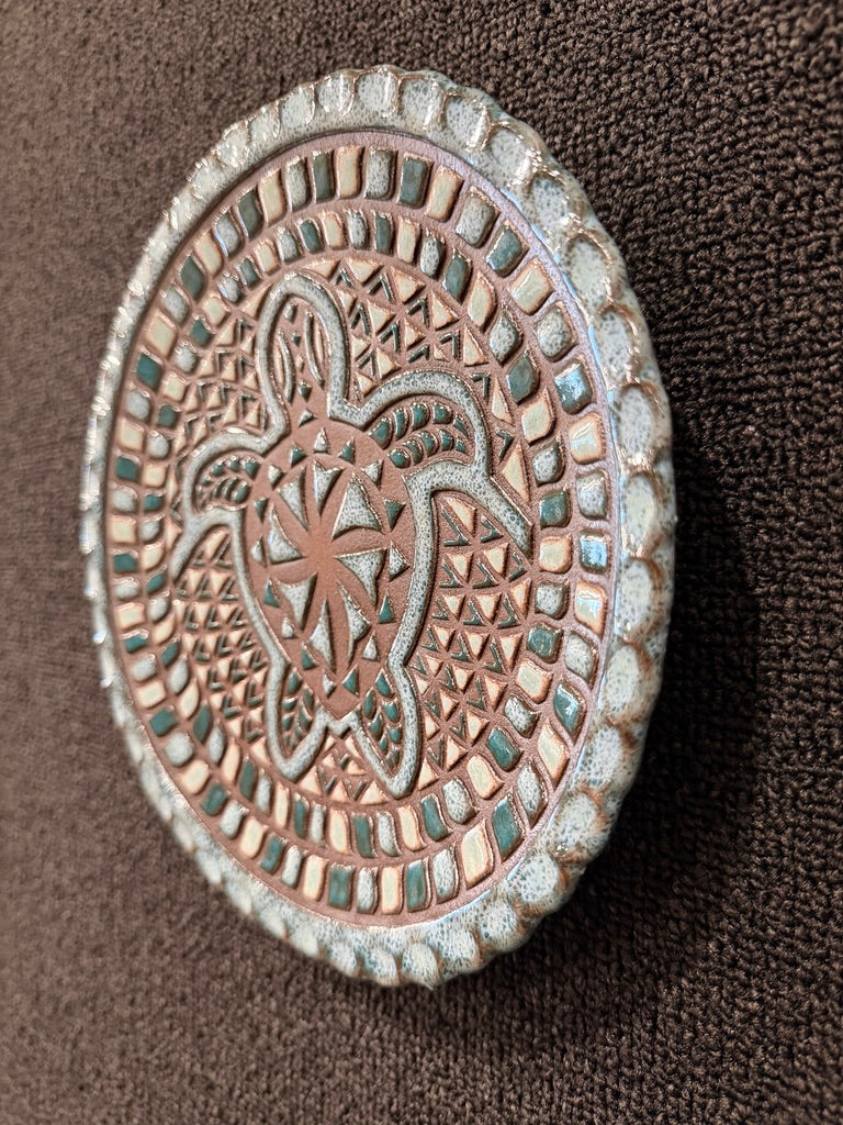 An oval decorative plaque or wall art piece with an embossed turtle and geometric pattern, featuring a weathered copper-brown center and a textured light gray border, displayed on a dark brown textured background.