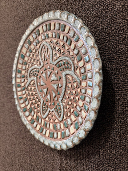 An oval decorative plaque or wall art piece with an embossed turtle and geometric pattern, featuring a weathered copper-brown center and a textured light gray border, displayed on a dark brown textured background.