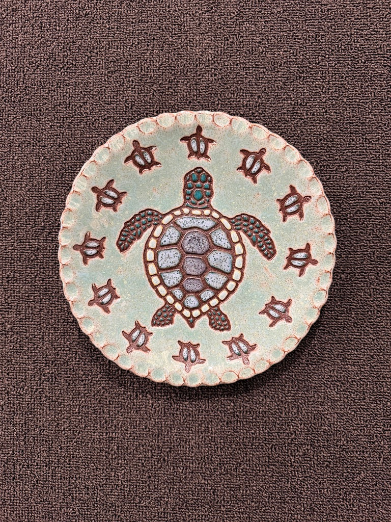 A round ceramic plate featuring a central sea turtle design with textured shell details and a border of small sea turtle motifs, set against a dark brown textured fabric background.