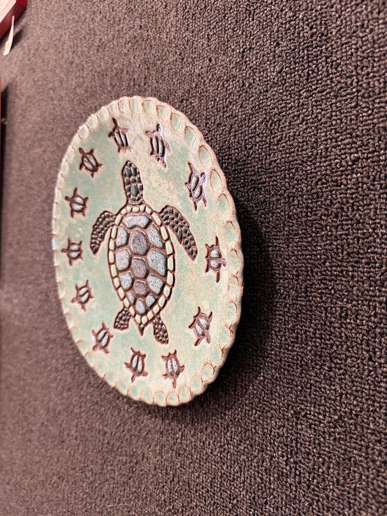 An oval ceramic decorative piece featuring a central sea turtle design surrounded by small star-like motifs, displayed on a textured dark brown surface.