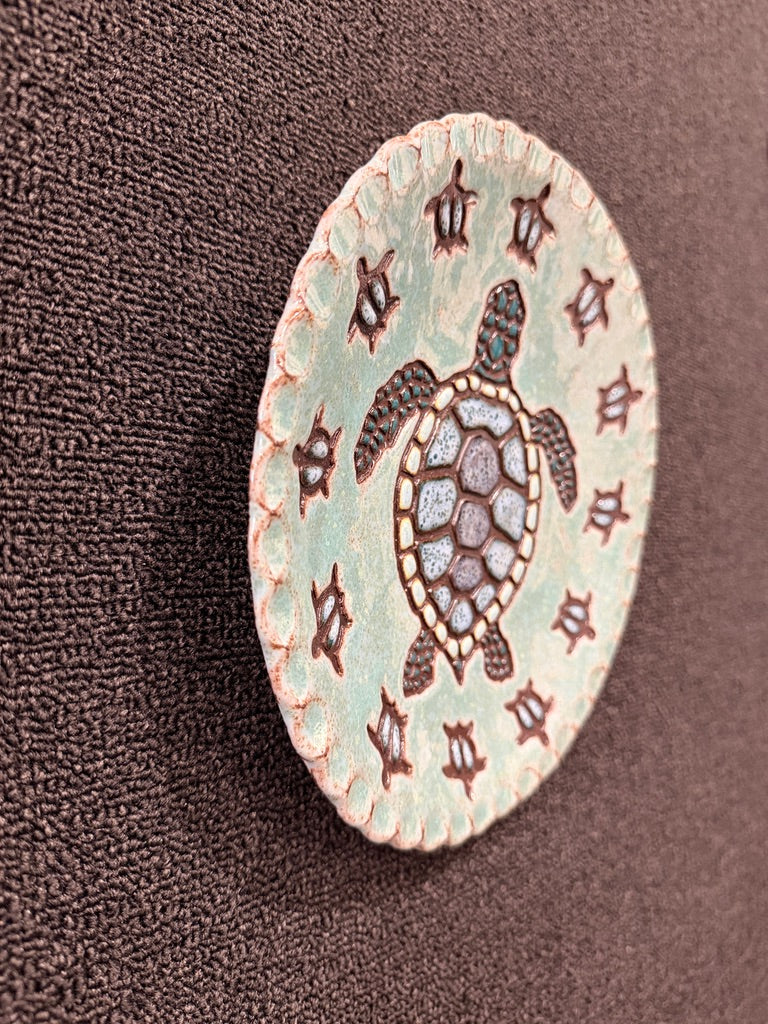 A round ceramic hanging plate featuring a central sea turtle design on a light mint green background, surrounded by a border of dark brown symbols, displayed on a textured dark brown fabric background.