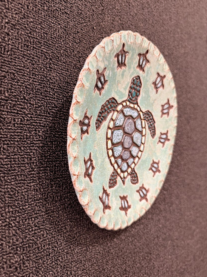 A round ceramic hanging plate featuring a central sea turtle design on a light mint green background, surrounded by a border of dark brown symbols, displayed on a textured dark brown fabric background.
