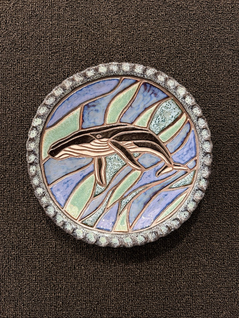 A round ceramic hanging plate featuring a stylized whale design with blue, green, and beige enamel sections, surrounded by a silver-tone beaded border on a dark brown textured background.