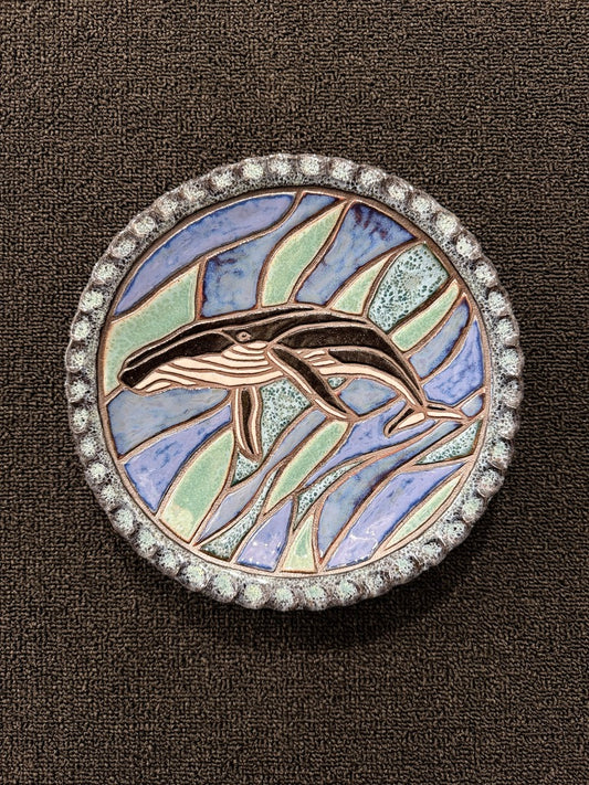 A round ceramic hanging plate featuring a stylized whale design with blue, green, and beige enamel sections, surrounded by a silver-tone beaded border on a dark brown textured background.