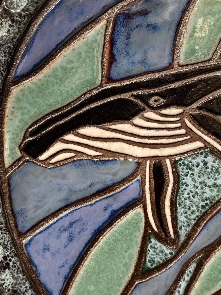 Close-up of a ceramic hanging plate design featuring a whale in cream and black, surrounded by blue, lavender, and green textured glass panels with dark brown outlines.