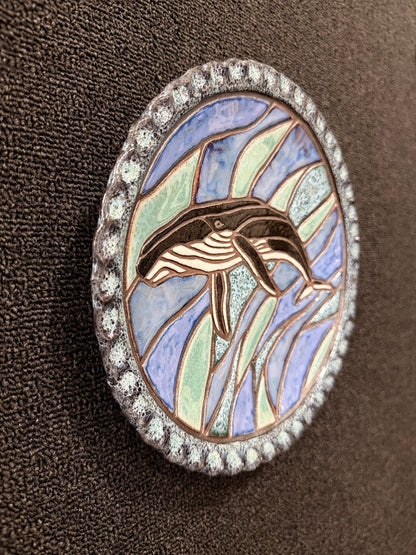 A side angle view of a round, ceramic  hanging plate featuring a whale design in the center, surrounded by blue and green mosaic-like segments, with a silver-toned beaded border, set against a dark brown textured background.