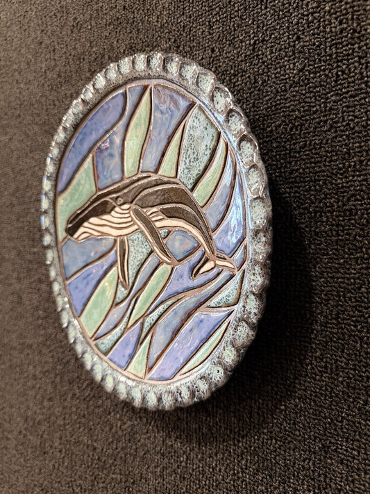 A side angle view of a round ceramic plate featuring a whale design over stylized wave-like segments in iridescent blue, green, and lavender tones, surrounded by a textured silver-toned border, shown on a dark brown textured background.